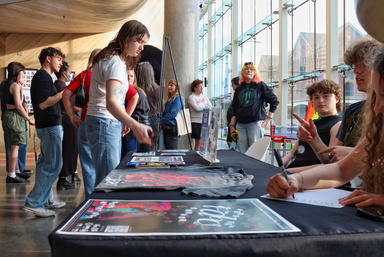 Students gather at a check-in table during the Iowa City Documentary Film Festival, with posters, merchandise, and volunteers interacting in a glass-walled lobby space.