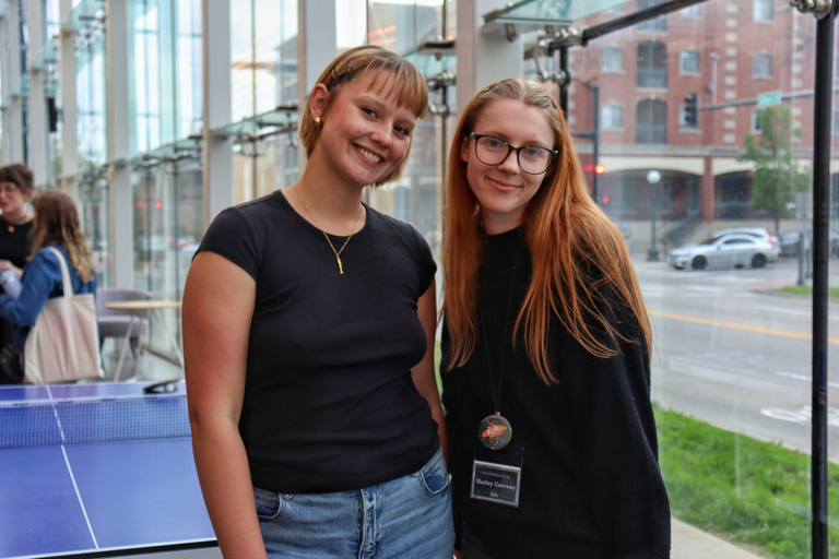 Two students stand together in a glass-walled lobby space.